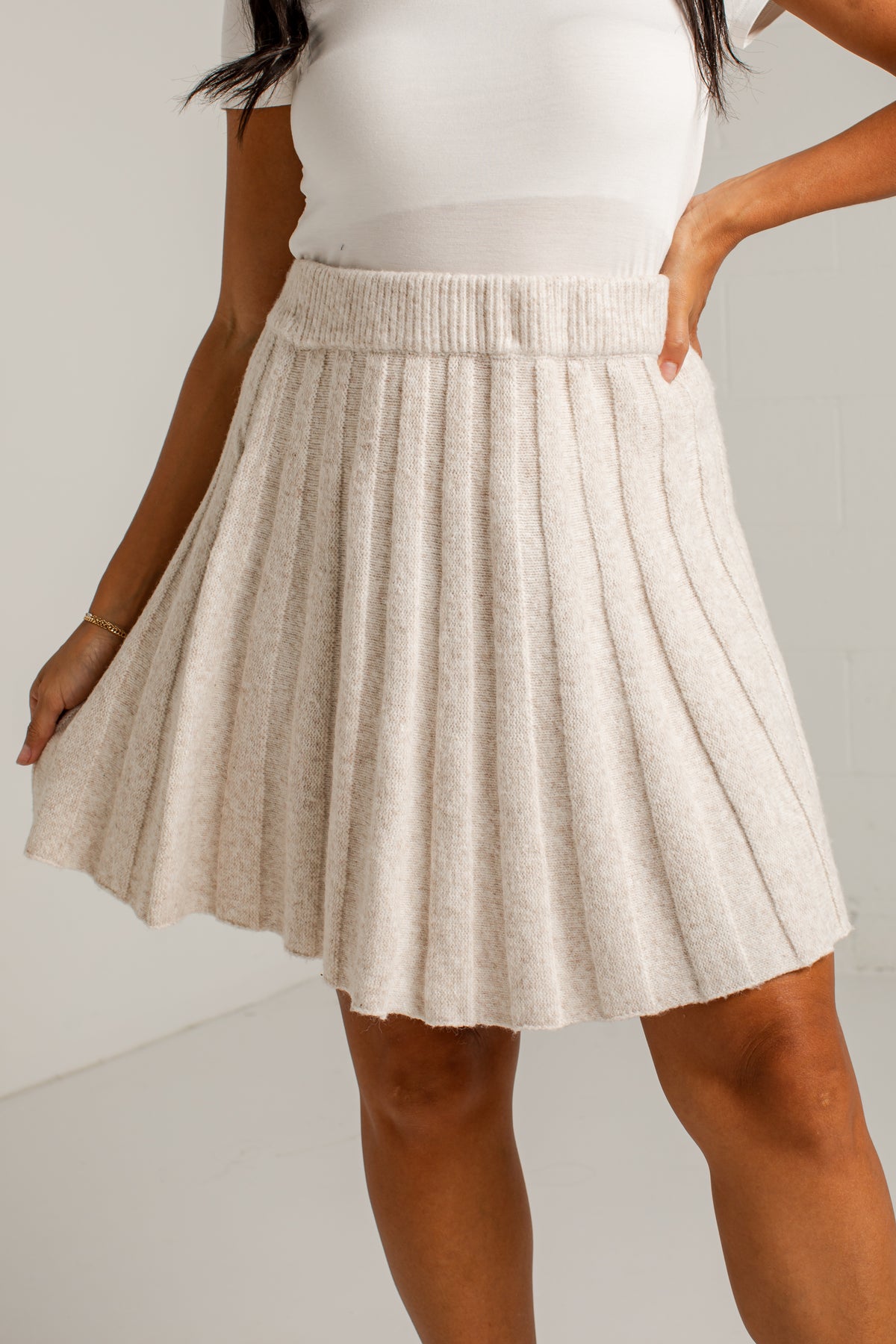 A woman wearing a cream-colored pleated knit mini skirt with a ribbed waistband, paired with a white top, shown from the chest down against a plain light background.