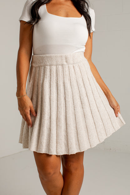 A woman wearing a white scoop-neck short-sleeve top and a pleated, textured oatmeal-beige mini skirt, standing against a light gray wall.