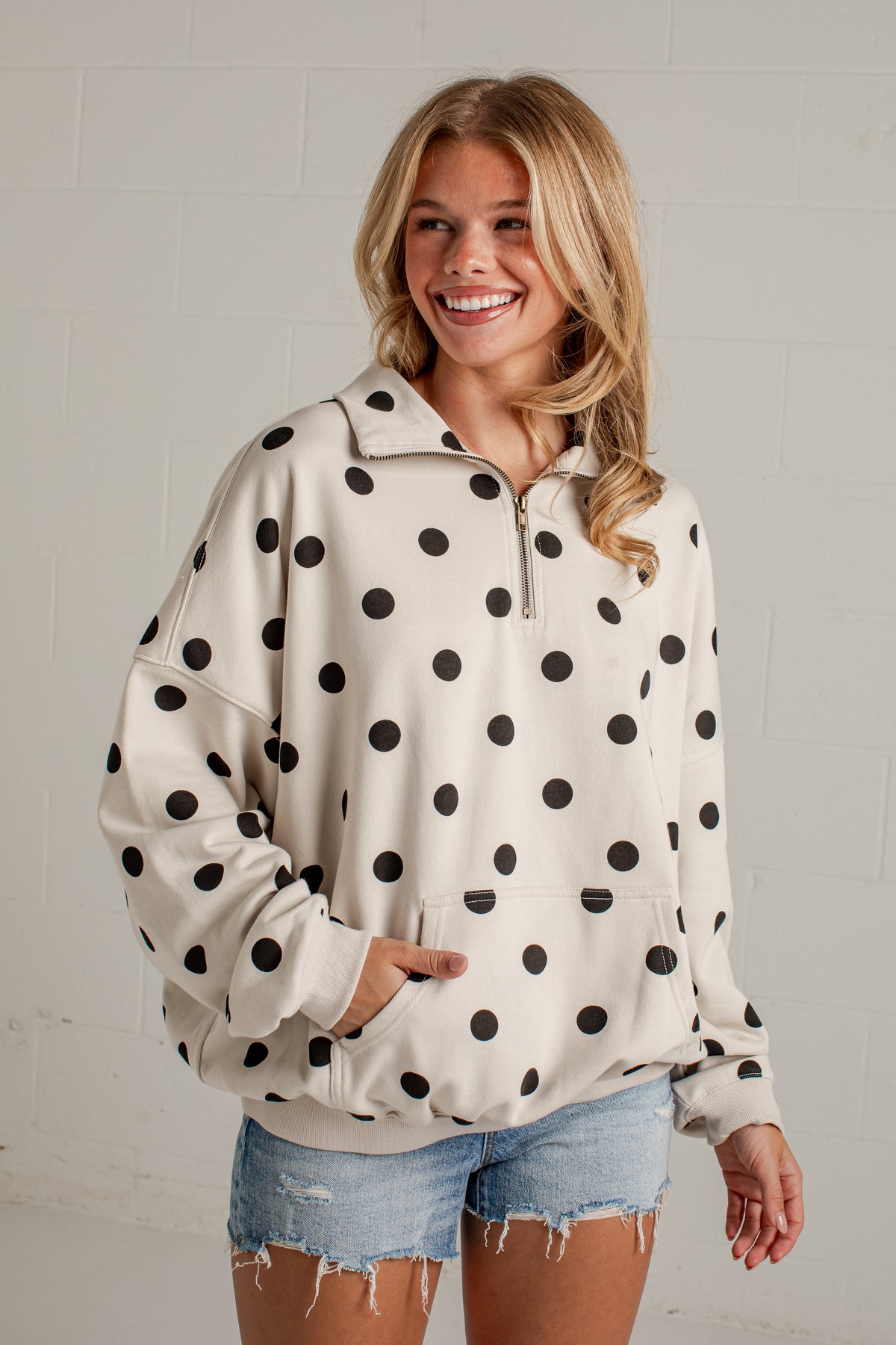 A woman wearing a cream-colored half-zip pullover sweatshirt with black polka dots, paired with light-wash denim shorts, standing against a white brick wall.