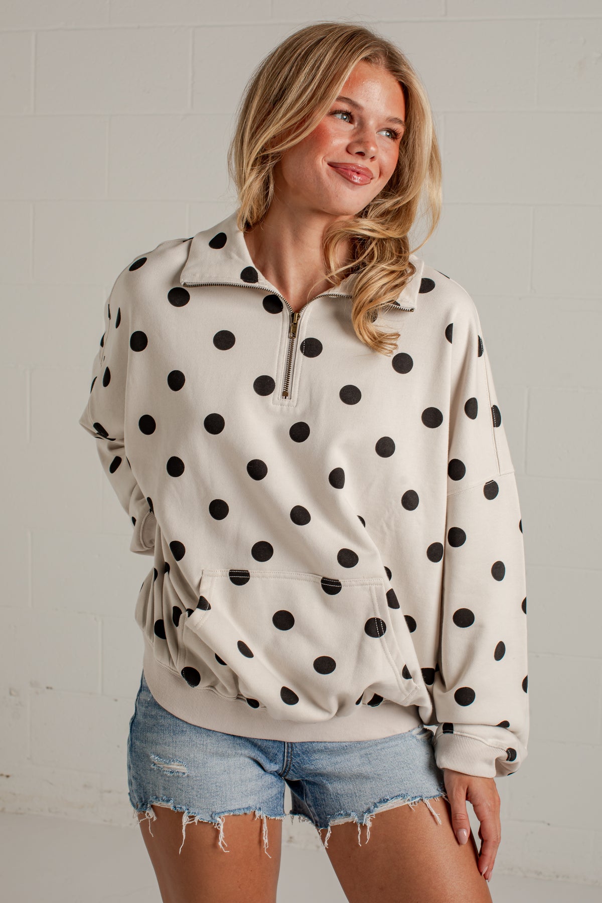 A woman wearing a cream-colored half-zip pullover sweatshirt with black polka dots, paired with distressed denim shorts, standing against a white brick wall.
