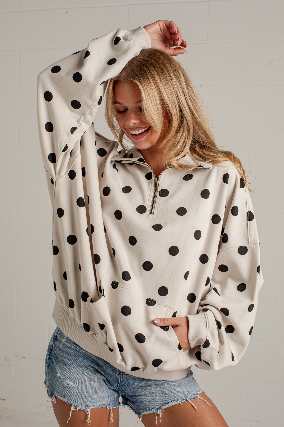 A woman wearing a cream-colored half-zip sweatshirt with black polka dots and denim shorts, posing against a light gray brick wall.