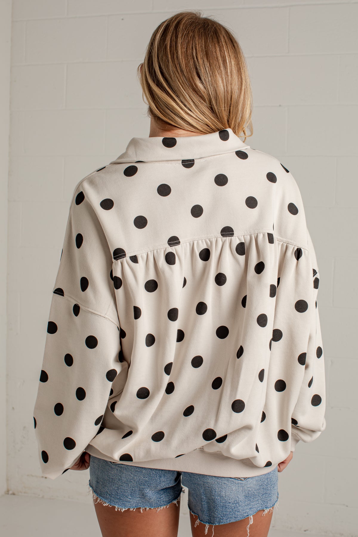 A woman viewed from behind wearing a cream-colored long-sleeve blouse with black polka dots and denim shorts, standing against a white brick wall.