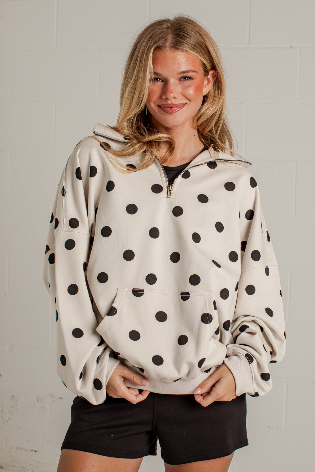 A woman wearing a cream-colored half-zip hoodie with black polka dots, paired with black shorts, standing against a light gray brick wall.