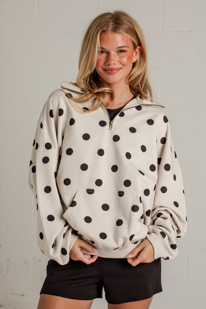A woman wearing a cream-colored half-zip hoodie with black polka dots, paired with black shorts, standing against a light gray brick wall.