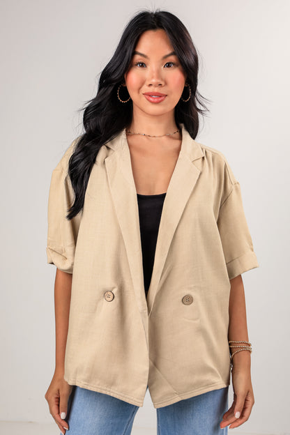 Woman wearing a beige blazer over a black top and blue jeans on a plain background