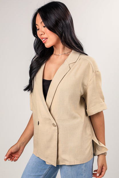 Woman wearing a beige blazer over a black top and blue jeans on a plain background
