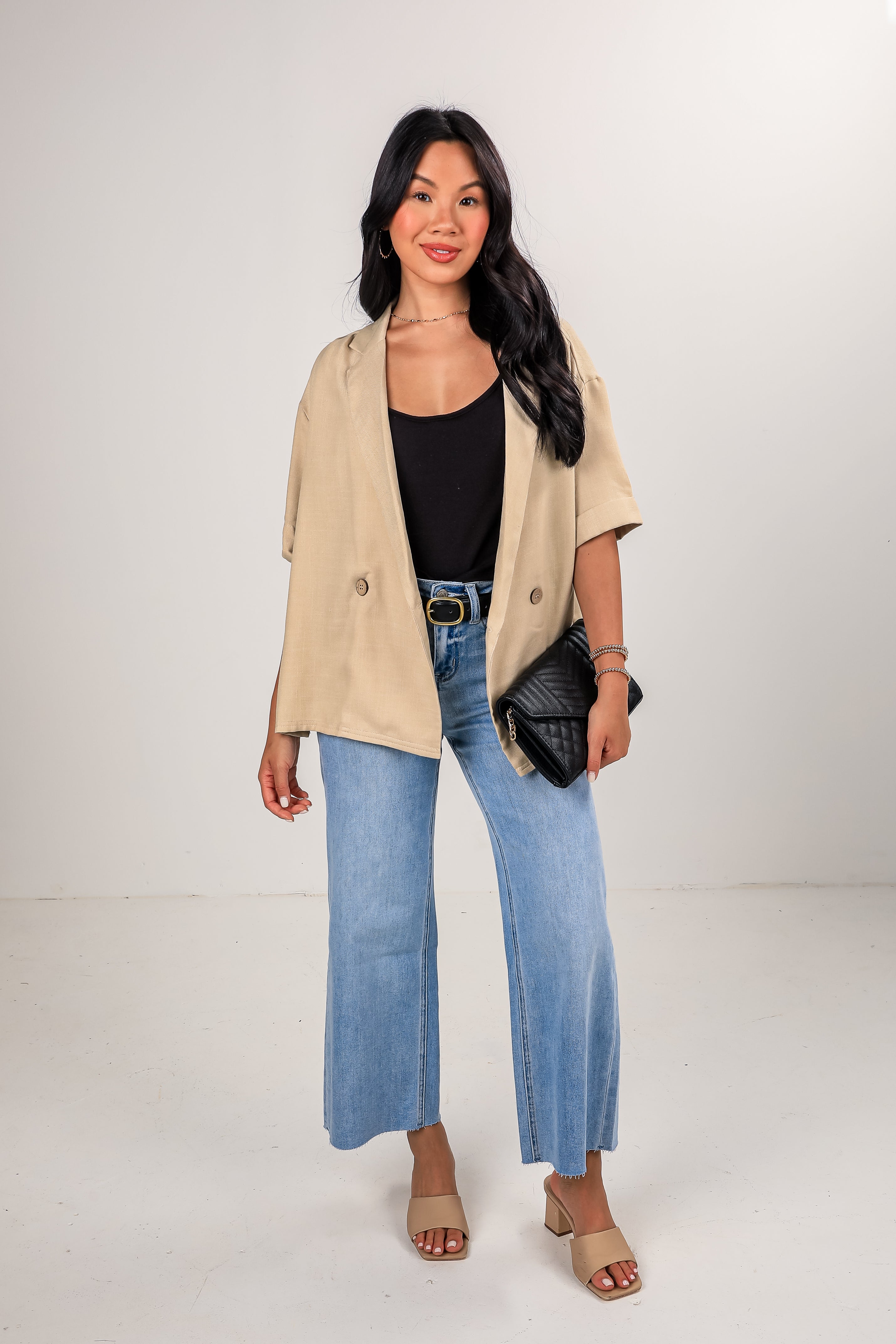 Woman wearing a beige jacket, black top, blue jeans, and sandals on a white background