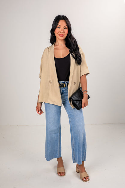 Woman wearing a beige jacket, black top, blue jeans, and sandals on a white background