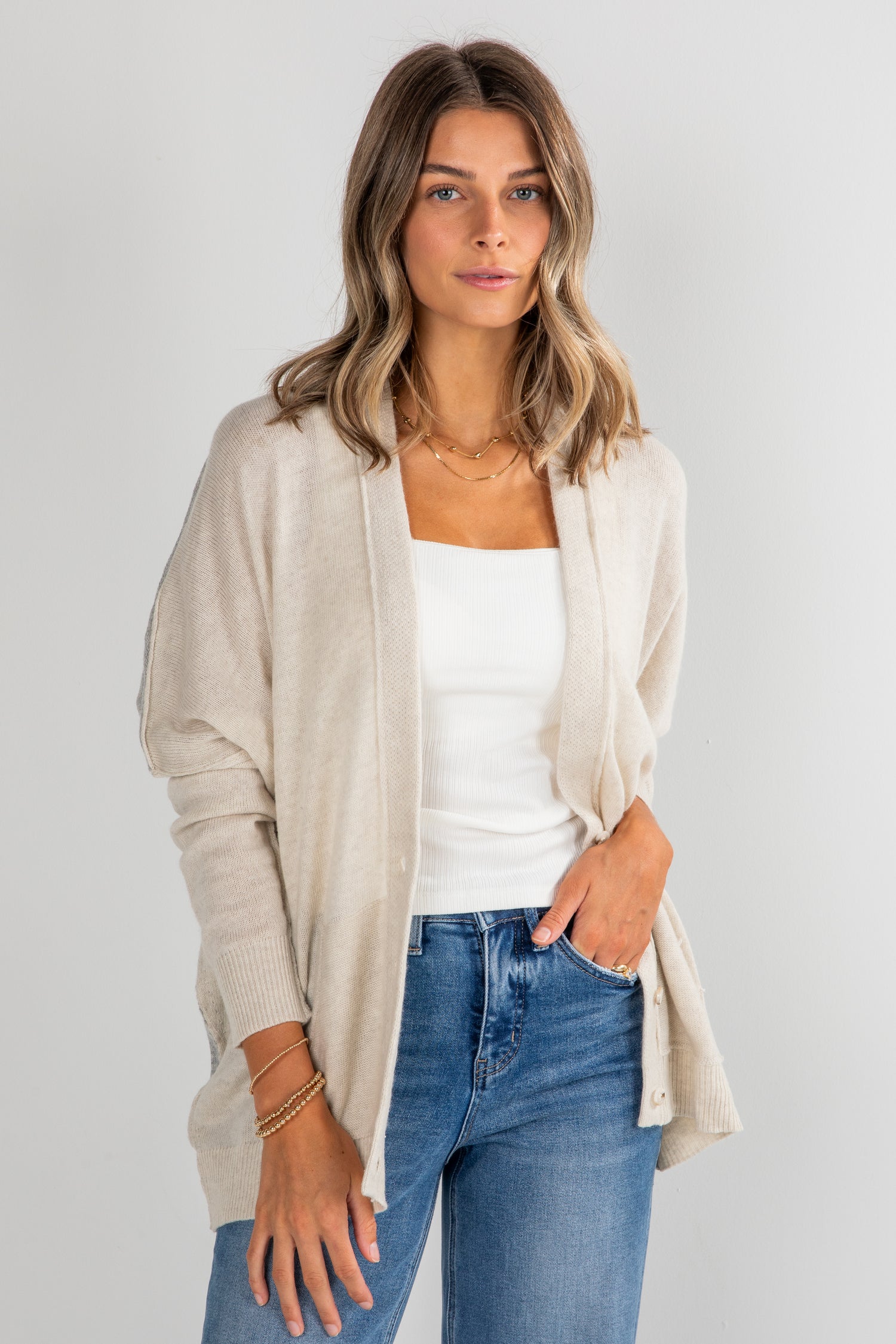 Woman wearing a beige cardigan, white top, and blue jeans on a plain background