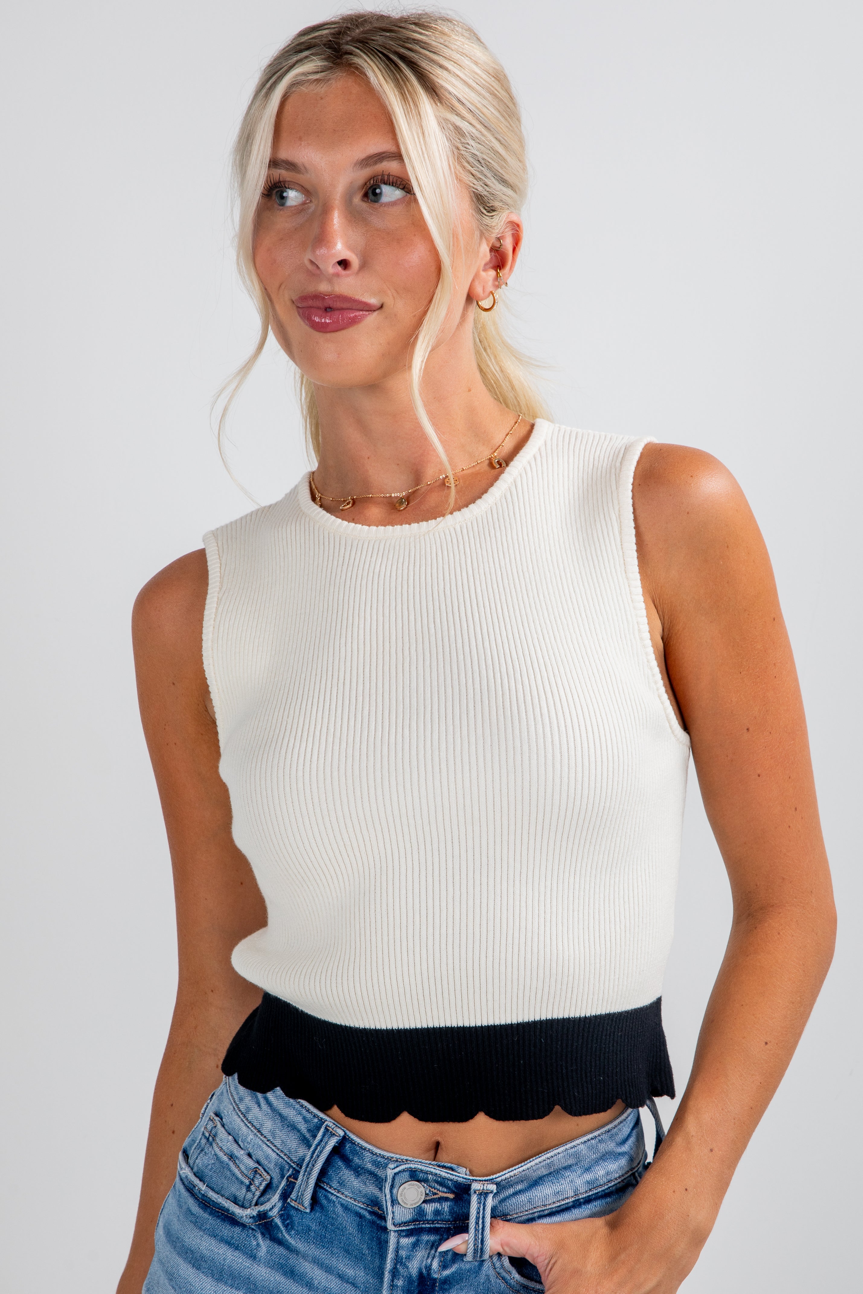 Woman wearing a white sleeveless top with black scalloped hem and blue jeans on a plain background