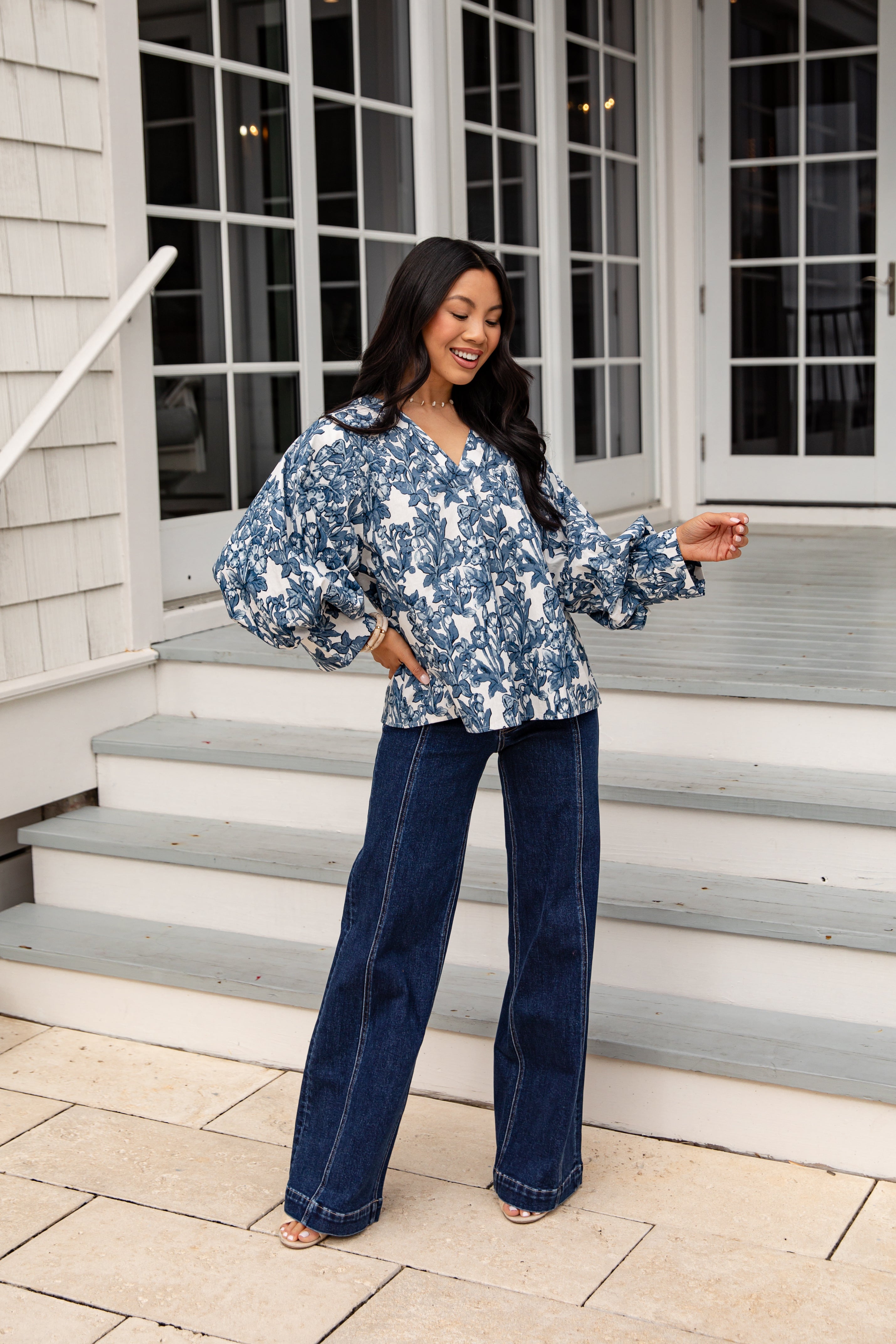 Woman in a blue floral blouse and jeans standing on steps outside a house.