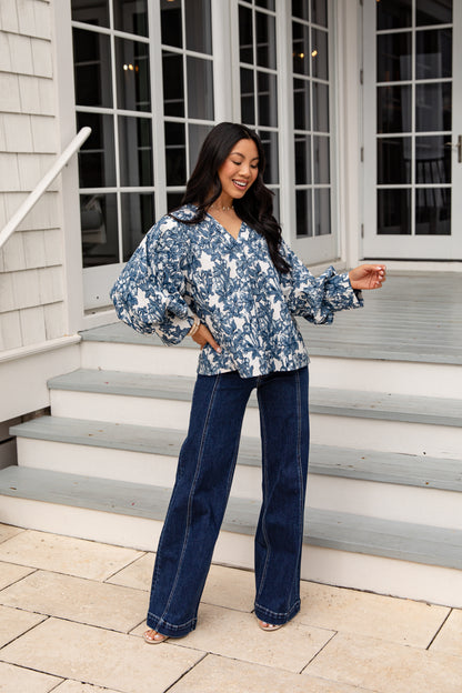 Woman in a blue floral blouse and jeans standing on steps outside a house.