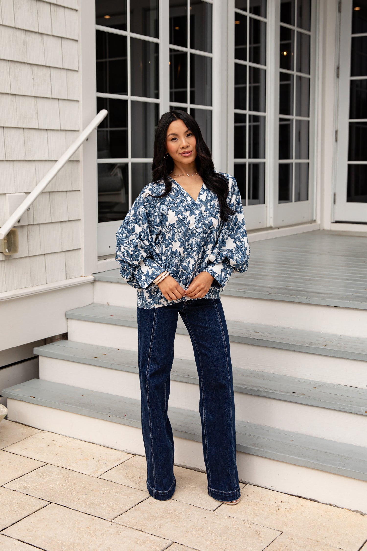Woman standing on a porch wearing a blue floral blouse and jeans.