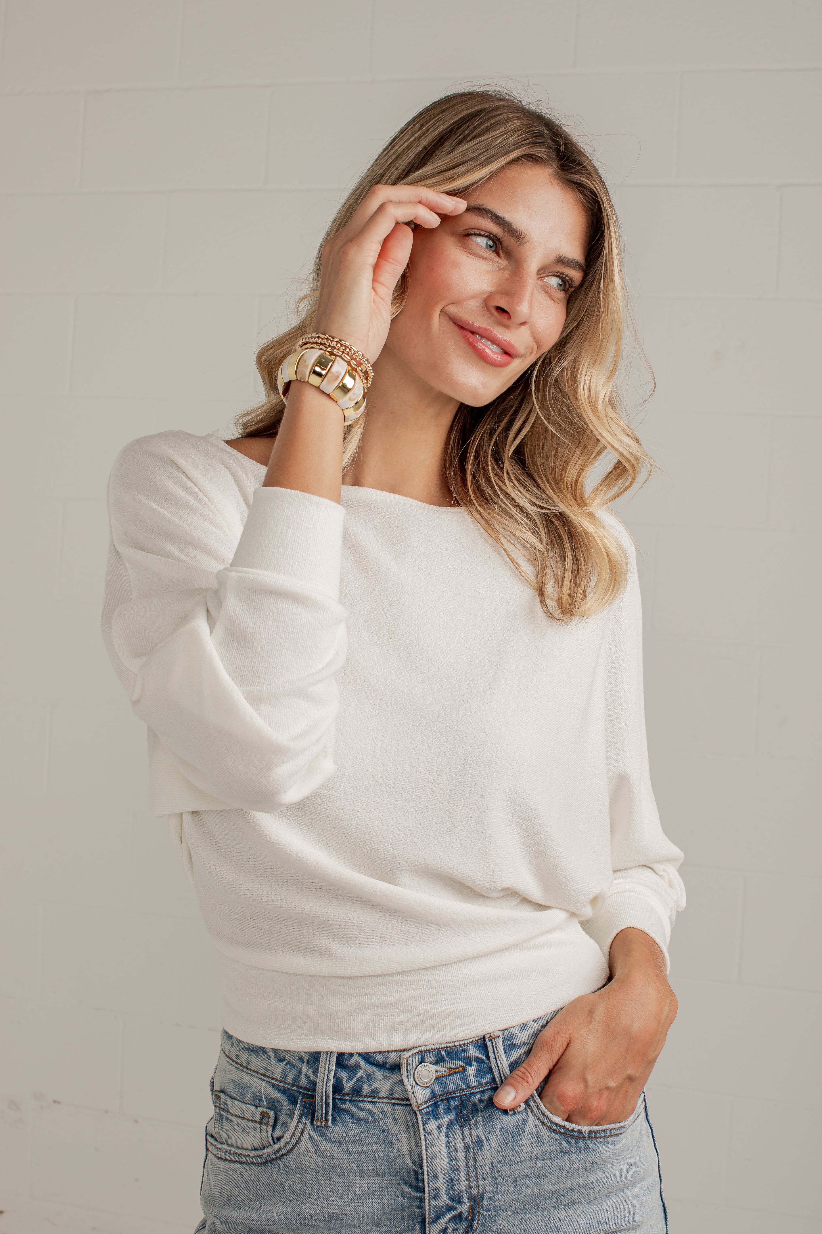 A woman wearing a white long-sleeve knit sweater and light-wash blue jeans, standing against a light gray brick wall.