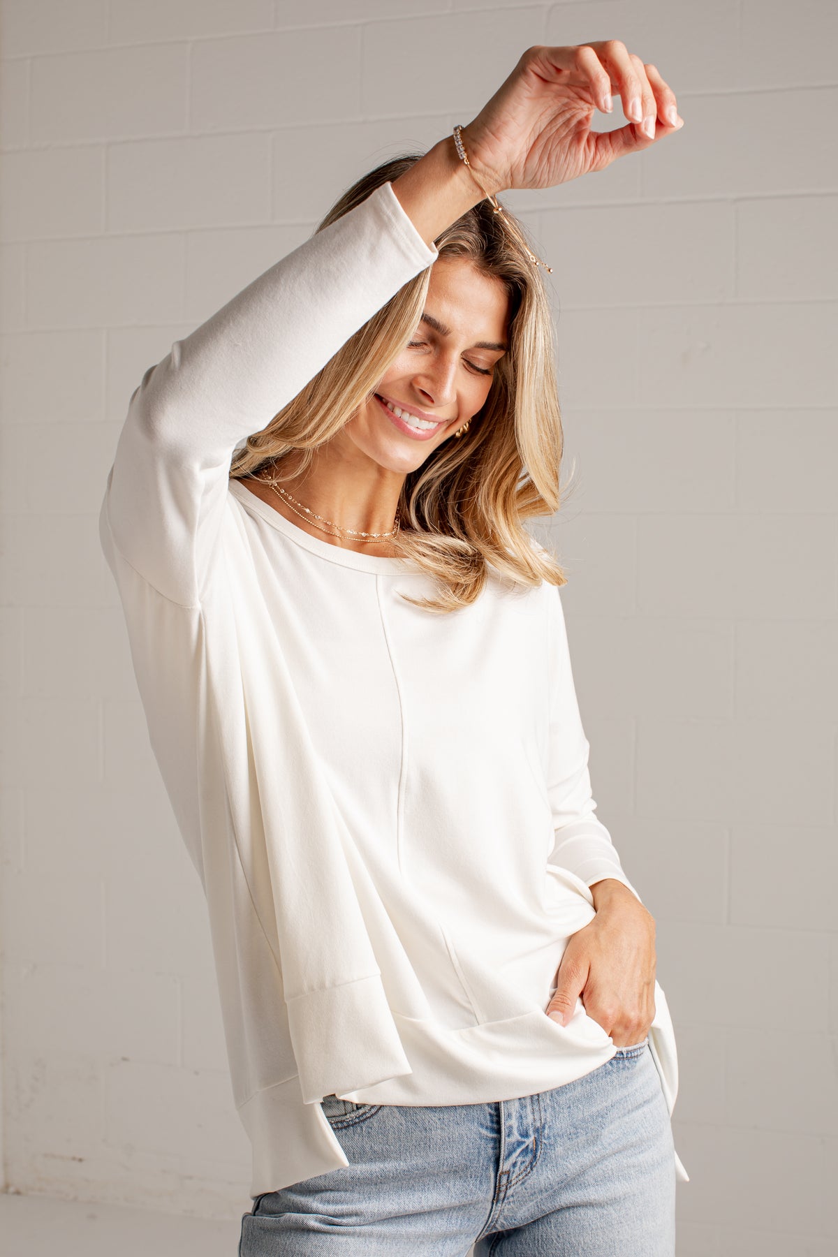 A woman wearing a loose-fitting, off-white long-sleeve top and light-wash blue jeans, posing against a light gray brick wall.