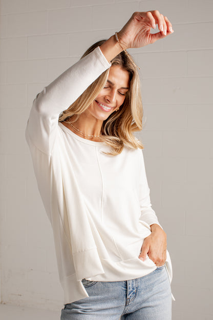 A woman wearing a loose-fitting, off-white long-sleeve top and light-wash blue jeans, posing against a light gray brick wall.