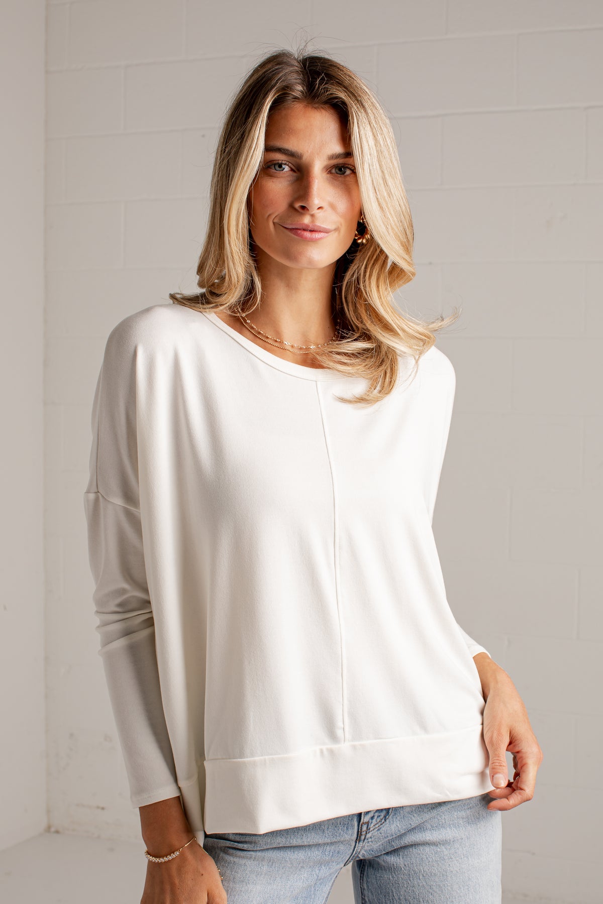 A woman with blonde hair wearing a cream-colored long-sleeve knit top and light blue jeans, standing against a white brick wall.