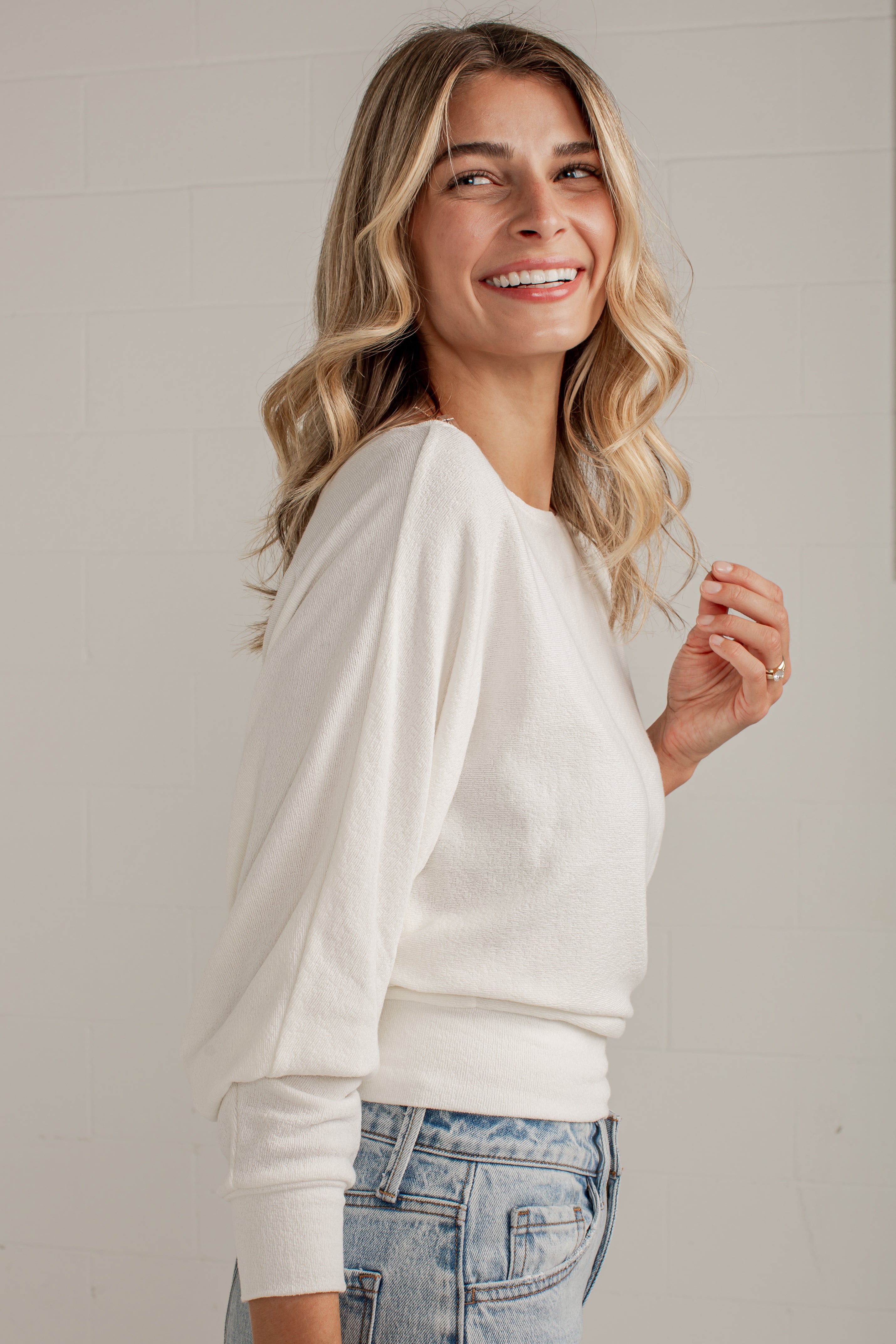 A woman with blonde wavy hair smiling and looking to the side, wearing a cream-colored long-sleeve knit top and light-wash blue jeans, standing against a light gray brick wall.