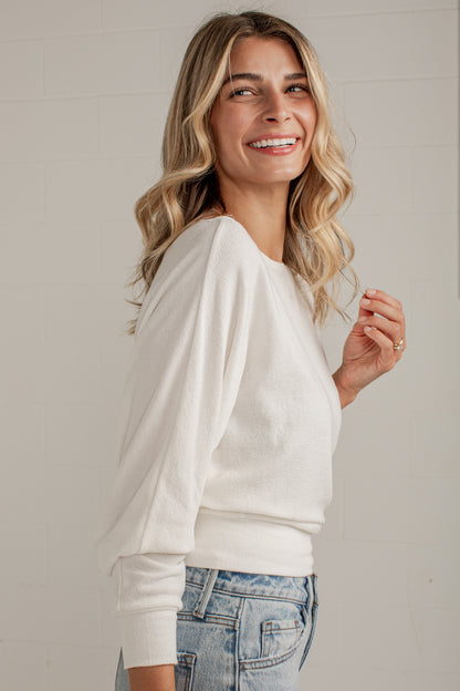 A woman with blonde wavy hair smiling and looking to the side, wearing a cream-colored long-sleeve knit top and light-wash blue jeans, standing against a light gray brick wall.