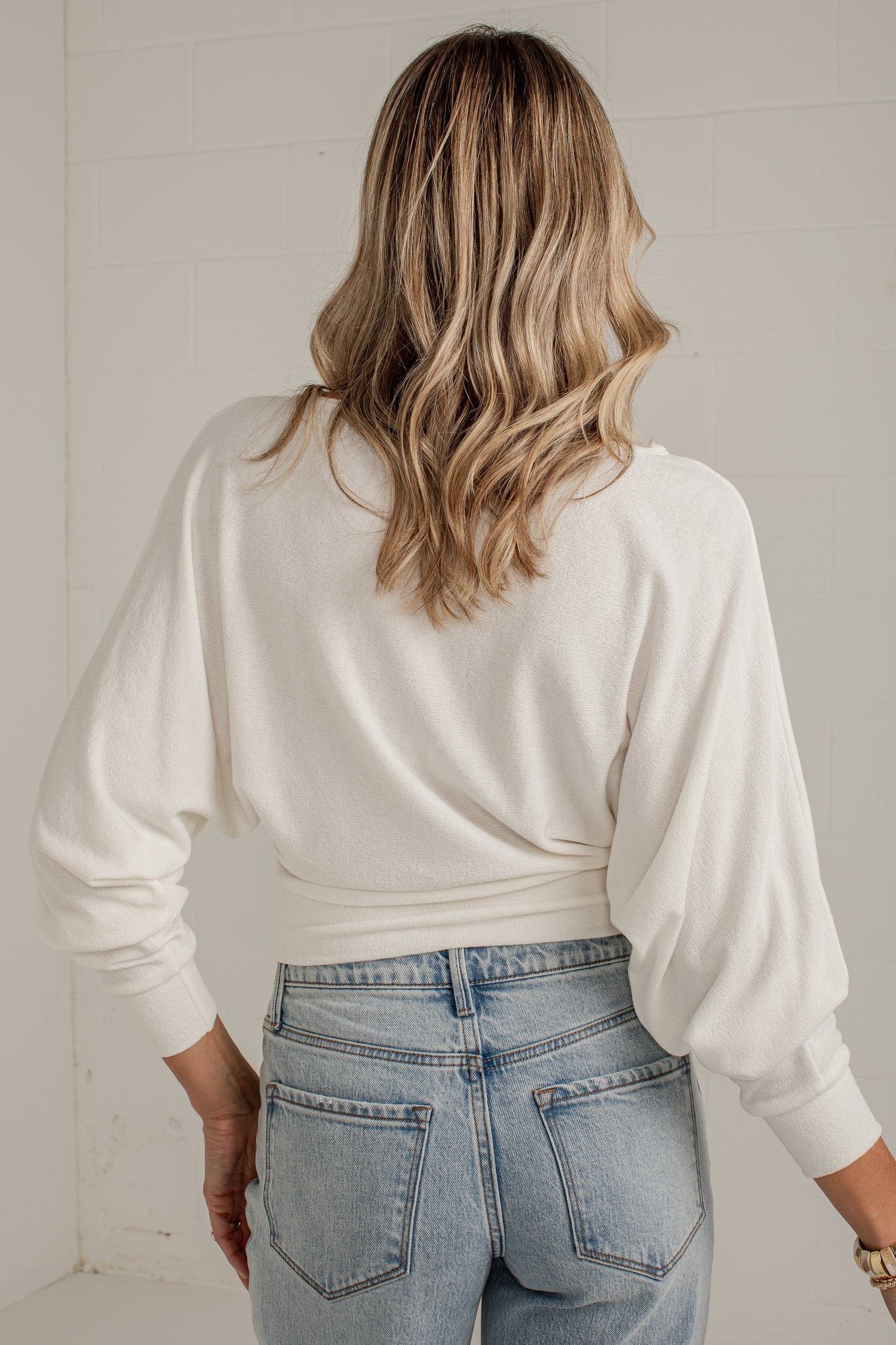 A woman viewed from behind wearing a cream-colored long-sleeve knit top and light-wash blue jeans, standing against a white tiled wall.