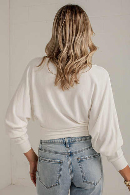 A woman viewed from behind wearing a cream-colored long-sleeve knit top and light-wash blue jeans, standing against a white tiled wall.