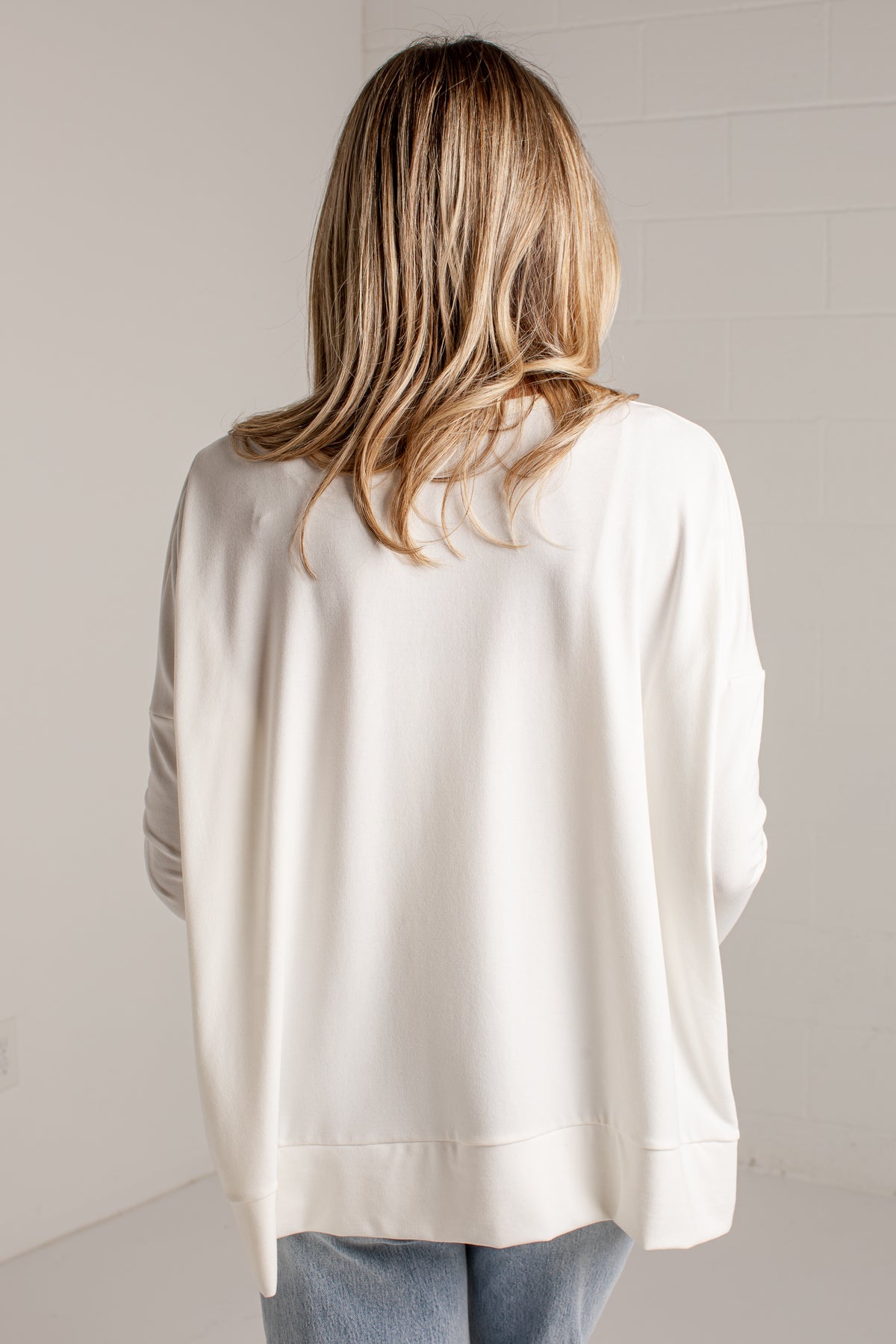 A woman viewed from behind wearing an oversized cream-colored long-sleeve top and light blue jeans, standing against a light gray wall.