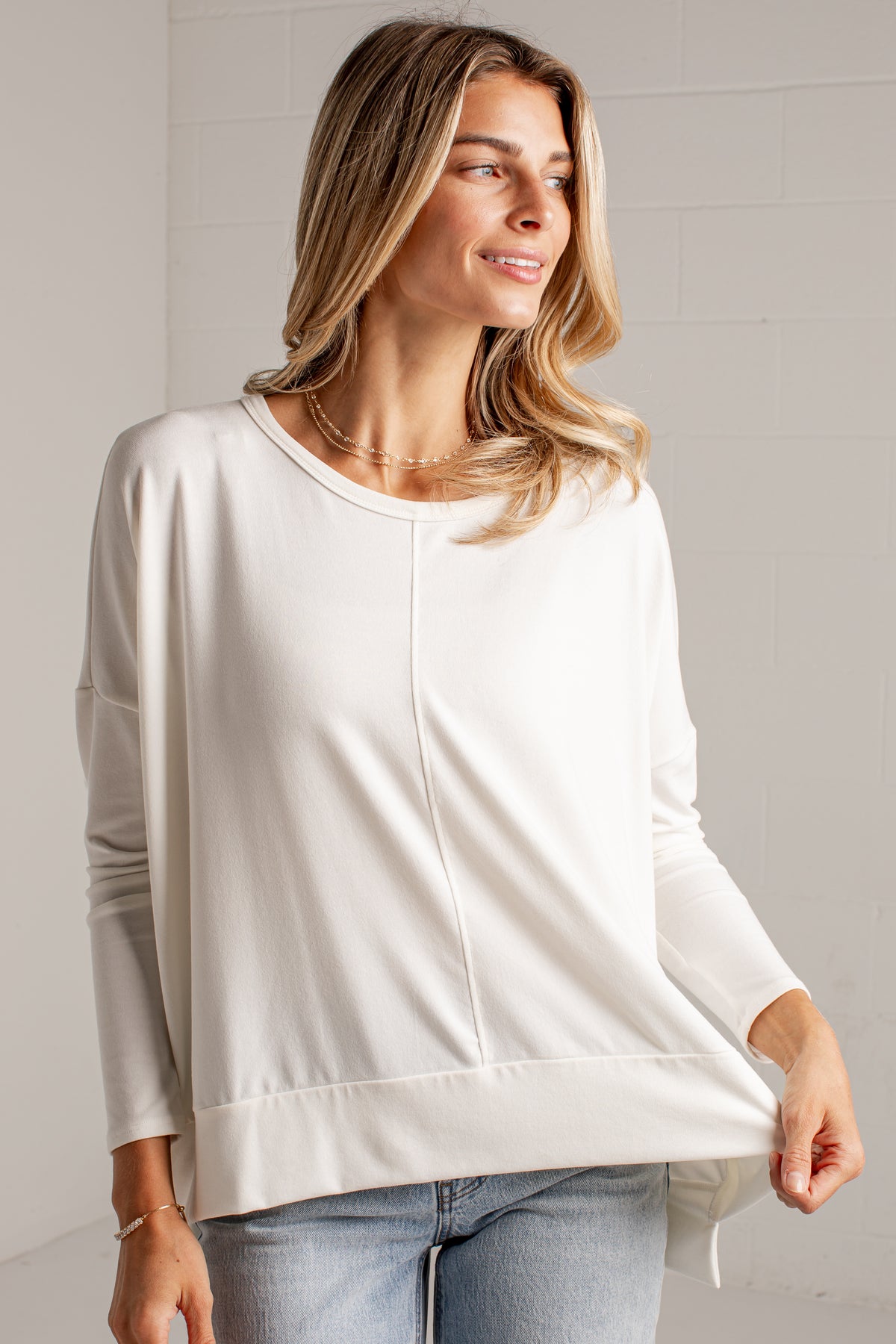 A woman wearing a white long-sleeve top with a relaxed, draped silhouette and a straight hem, paired with light-wash blue jeans, standing against a white brick wall.