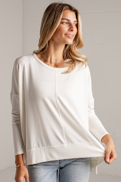 A woman wearing a white long-sleeve top with a relaxed, draped silhouette and a straight hem, paired with light-wash blue jeans, standing against a white brick wall.