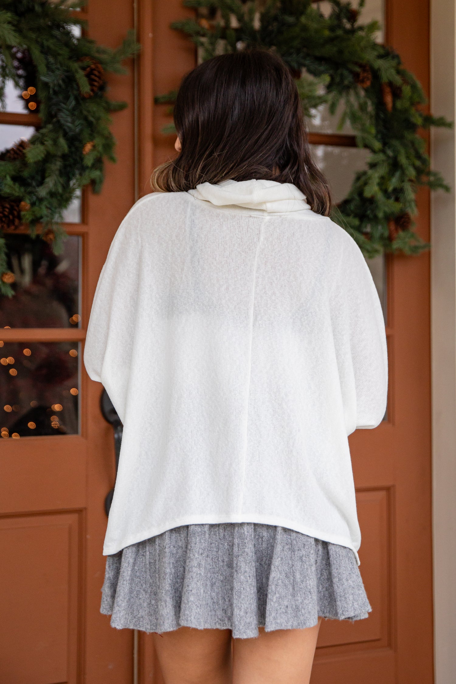 Person wearing a white sweater and gray skirt in front of a wooden door with greenery.