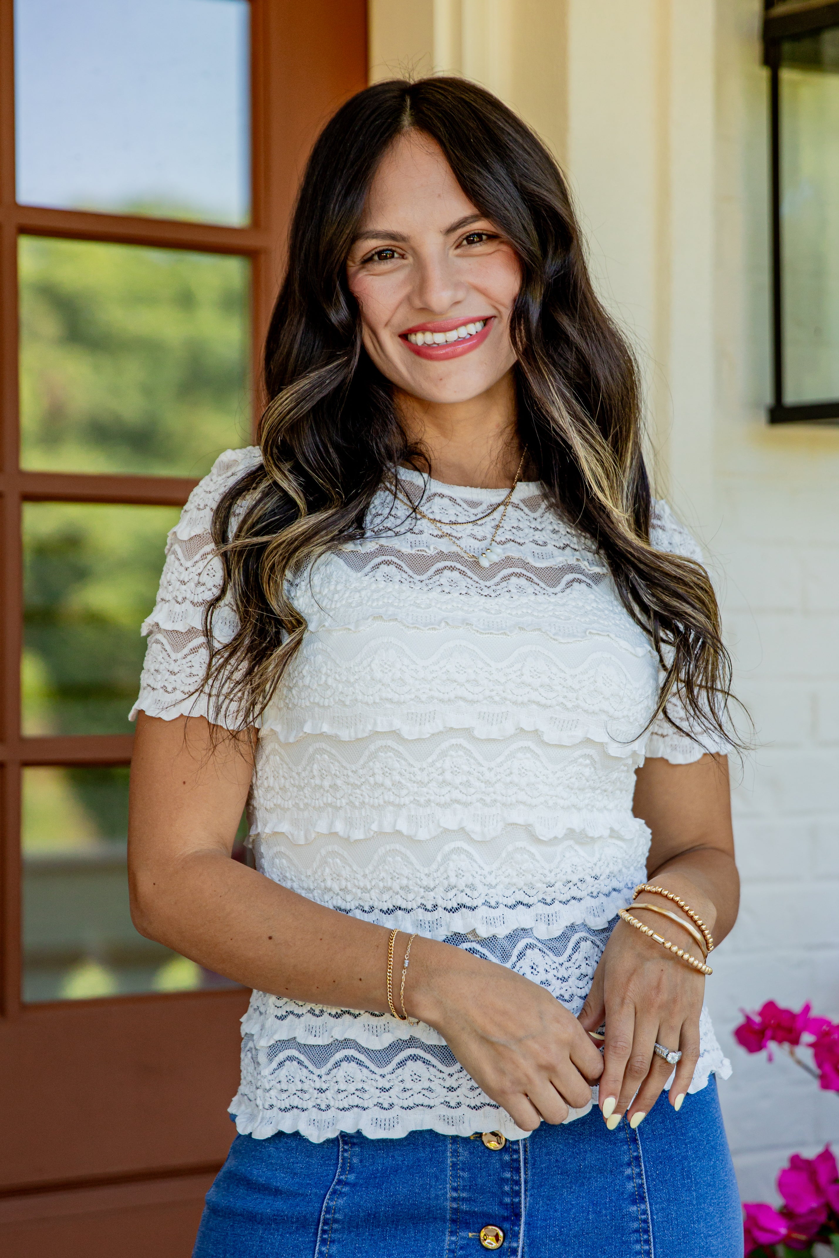 A woman smiling and wearing a white short-sleeve lace top with ruffle details, paired with blue jeans, standing in front of a porch with a brown door and white brick wall.