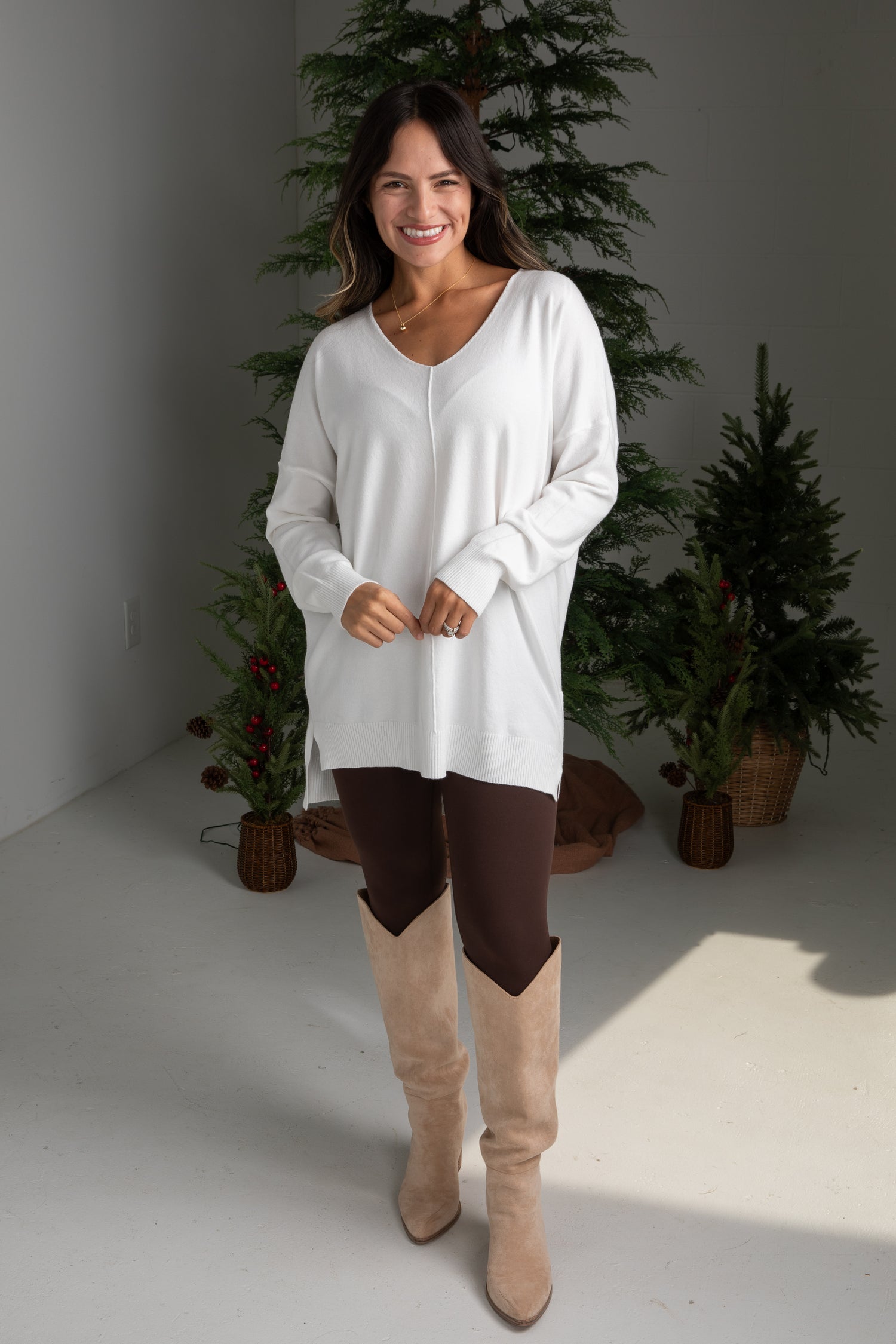 Woman wearing a white long-sleeve top and brown pants with beige boots, standing in front of decorative plants.