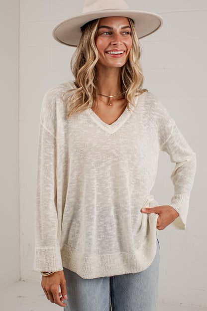 A woman wearing a cream-colored, long-sleeve V-neck knit sweater, light-wash jeans, and a wide-brimmed beige hat, standing against a white paneled wall.