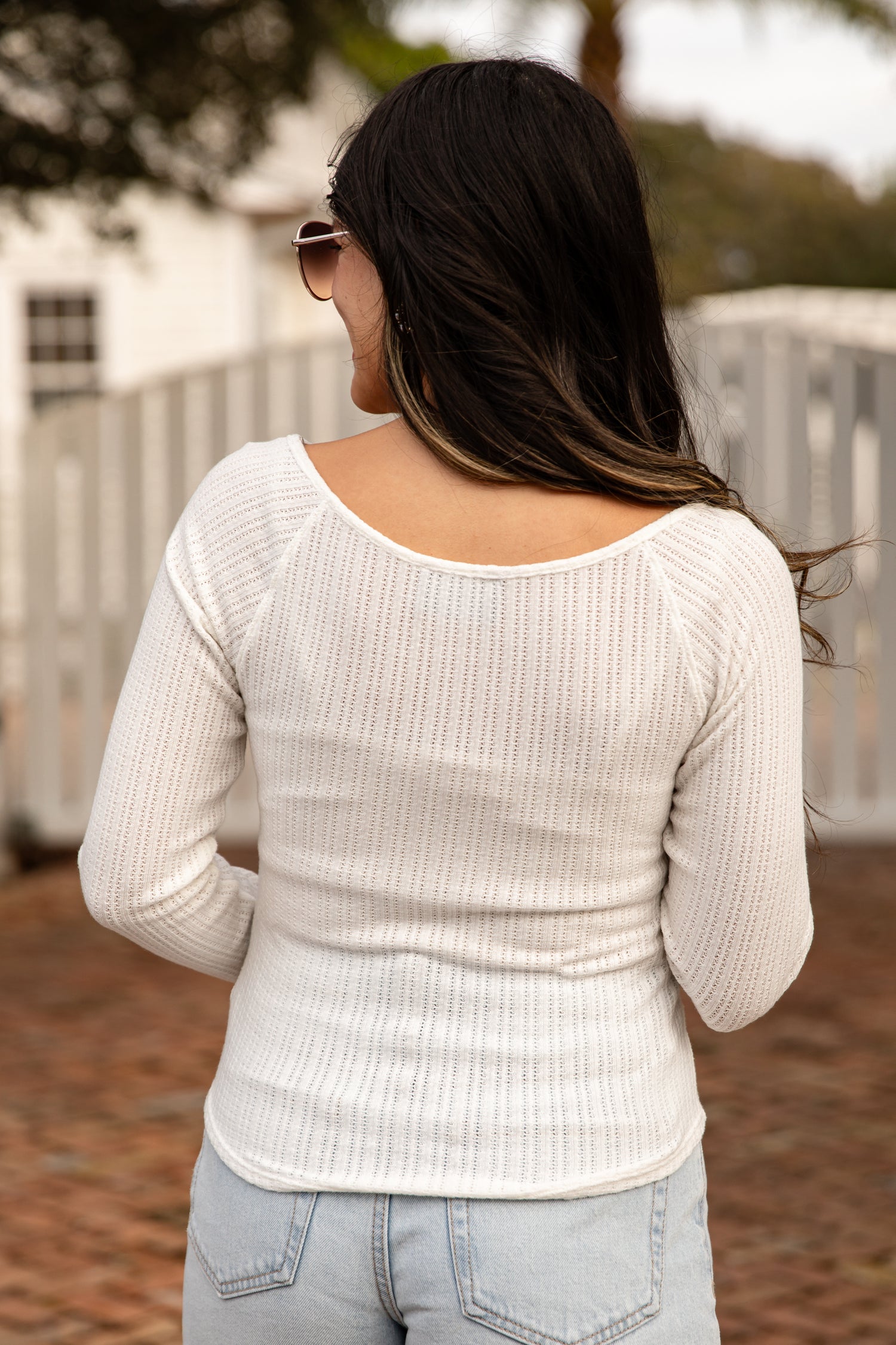Woman wearing a beige sweater and sunglasses, standing outdoors with trees and a fence in the background.