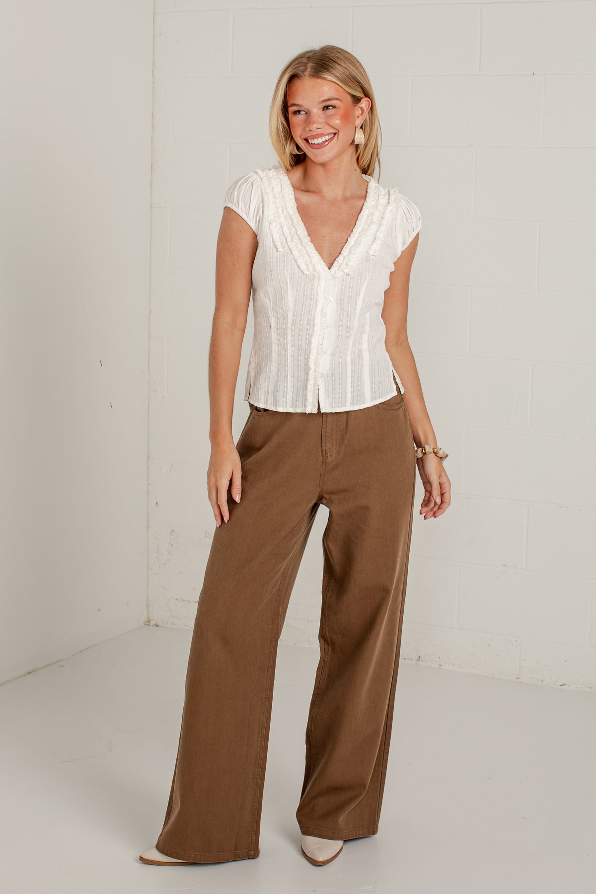 A woman modeling a white short-sleeve button-front blouse with pleated detailing and wide-leg brown pants, standing against a white studio wall.