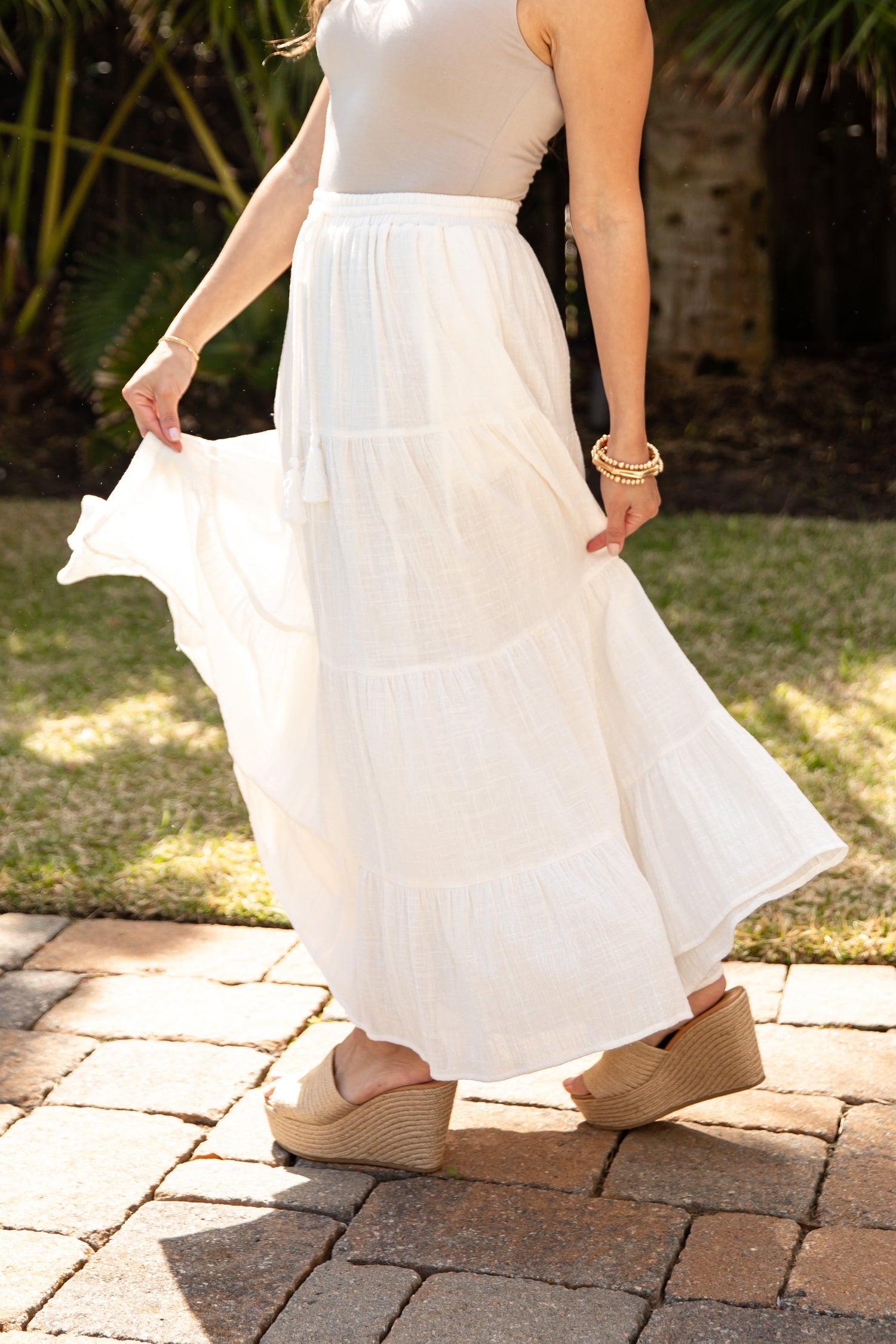 Person wearing a white skirt and beige sandals on a paved area with grass and plants in the background