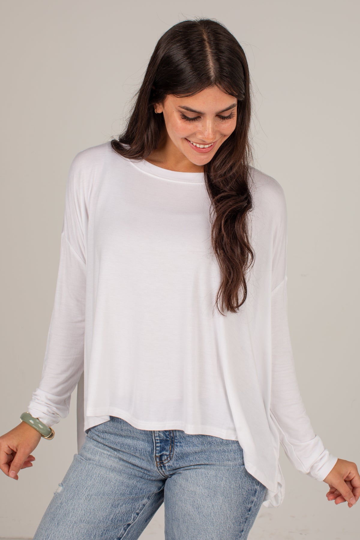 A woman wearing a white long-sleeve crewneck top and light blue jeans, standing against a plain light gray background.