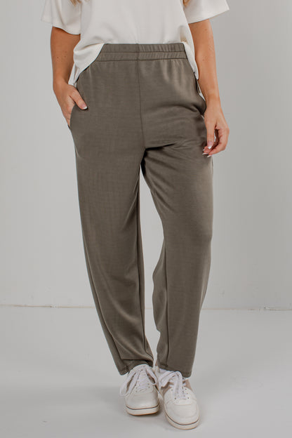A person wearing loose, straight-leg jogger pants in a muted taupe-gray color, paired with a white t-shirt and white sneakers, standing against a light gray studio background.