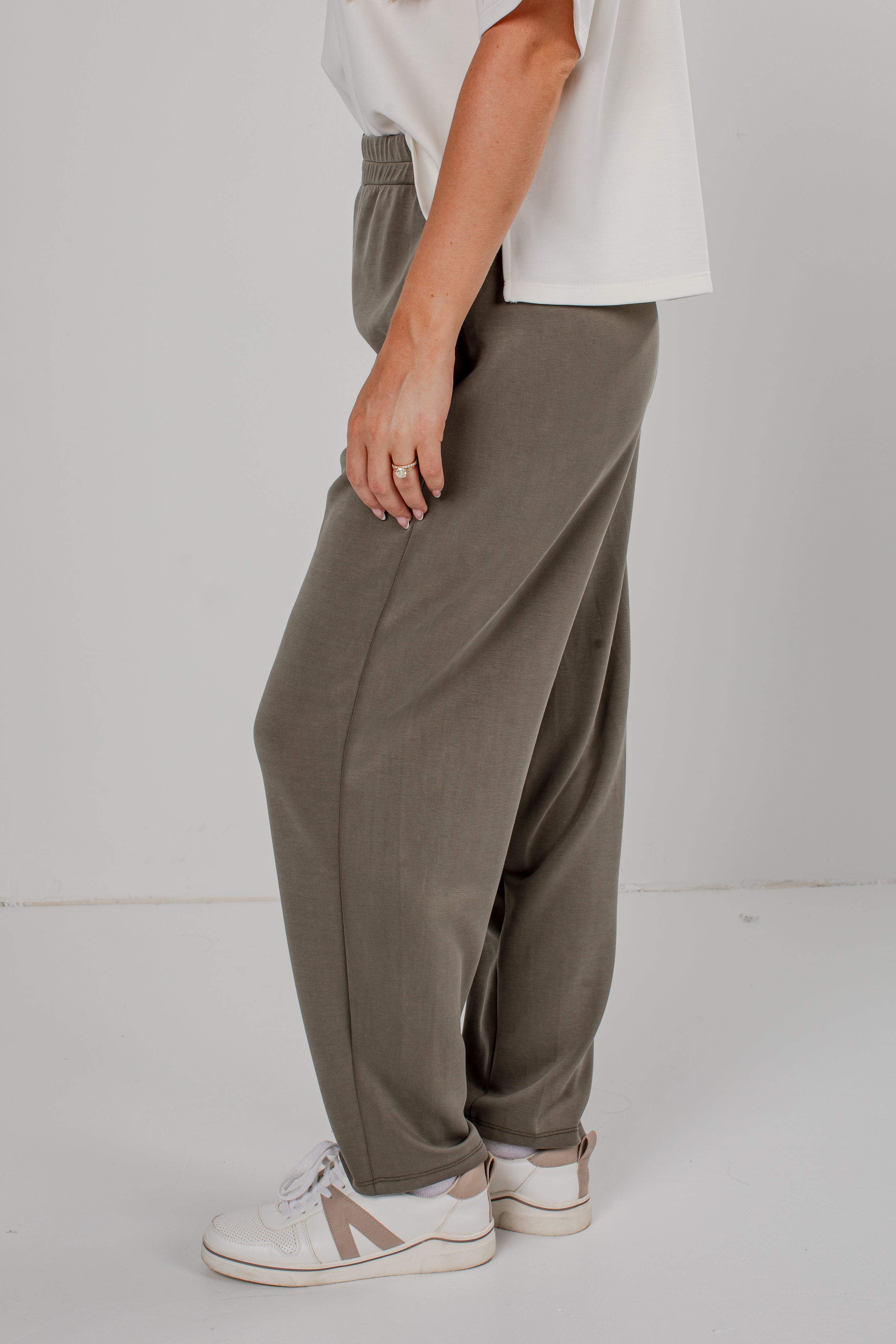 A person wearing loose, wide-leg taupe pants with an elastic waistband, paired with a white t-shirt and white sneakers, shown from the waist down against a light gray studio background.