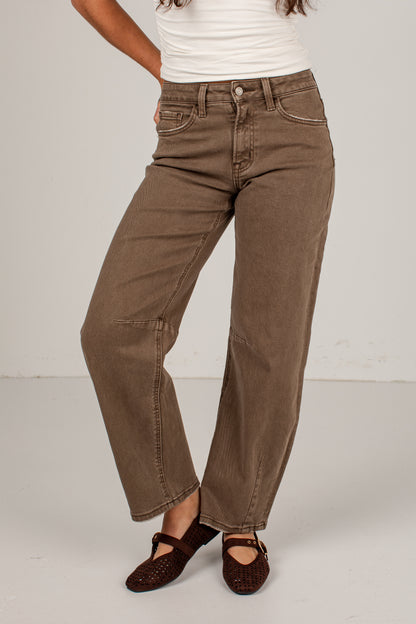 A person wearing brown wide-leg cropped jeans with a white ribbed top and dark brown Mary Jane flats, standing against a light gray studio background.