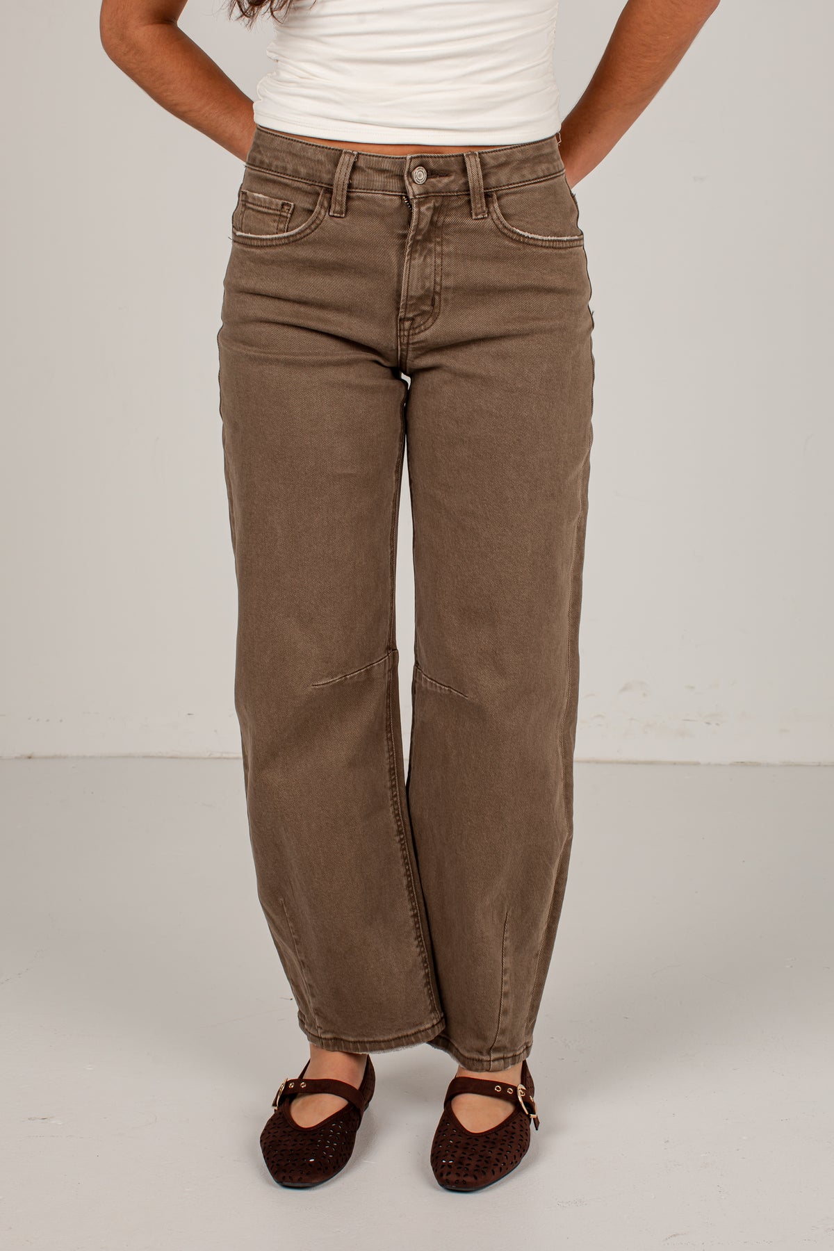 A person wearing brown wide-leg cropped jeans with a white top and dark brown glittery Mary Jane shoes, standing against a light gray studio background.
