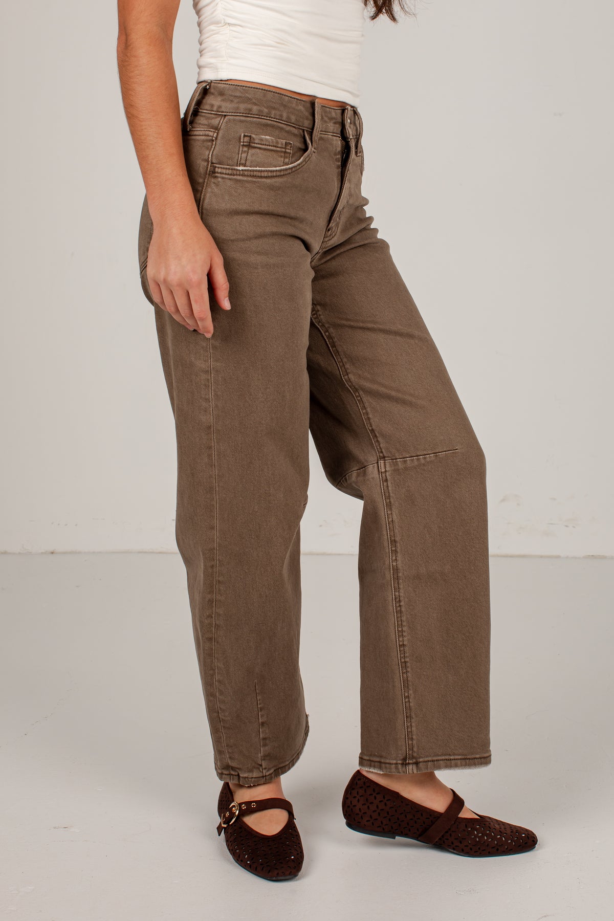 A person wearing brown wide-leg jeans with a white top and dark brown flat shoes, standing against a plain light gray studio background.