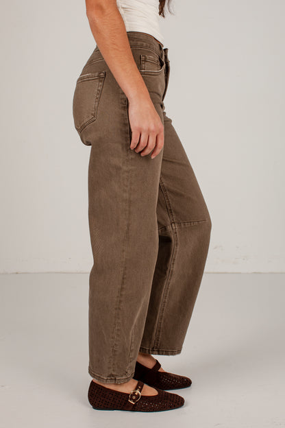 A side-profile view of a person wearing brown wide-leg cropped jeans, a white top, and dark brown woven flats against a plain light gray studio background.