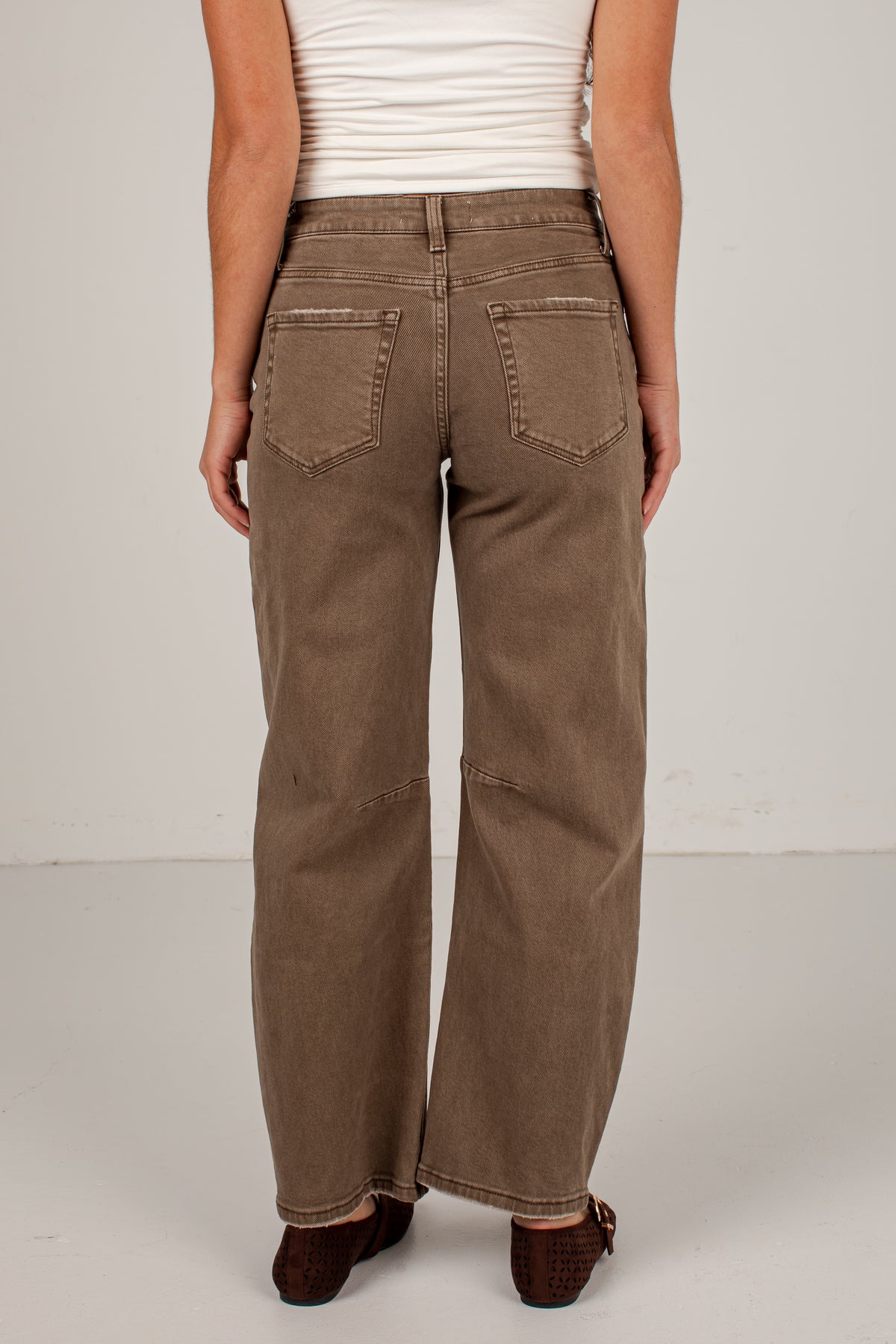 A person shown from the back wearing wide-leg brown jeans, a white ribbed tank top, and dark brown flat shoes against a plain light gray studio background.