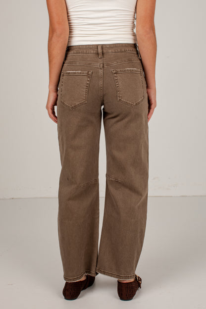 A person shown from the back wearing wide-leg brown jeans, a white ribbed tank top, and dark brown flat shoes against a plain light gray studio background.
