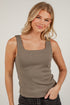 A woman wearing a fitted sleeveless tank top in a muted taupe-brown color, paired with dark jeans, standing against a white brick wall.