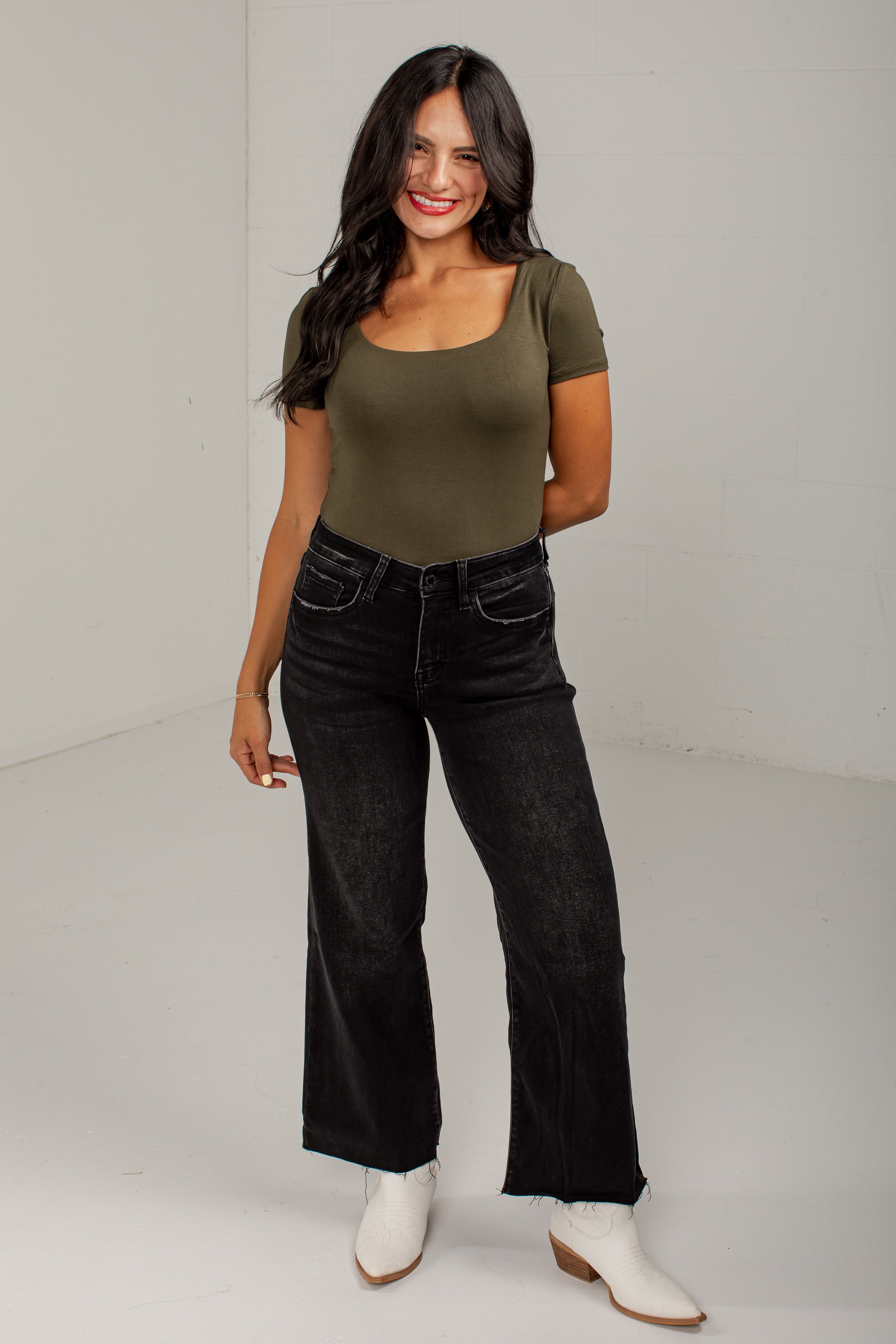 A woman modeling a fitted short-sleeve scoop-neck top in olive green, paired with black wide-leg jeans and white ankle boots, standing against a plain light gray studio background.