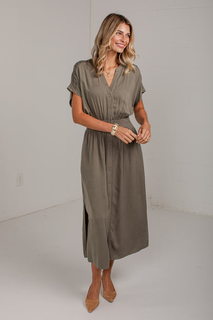 A woman modeling a short-sleeve, button-front midi dress in a muted olive-gray or taupe-brown shade, standing against a plain light gray studio wall.