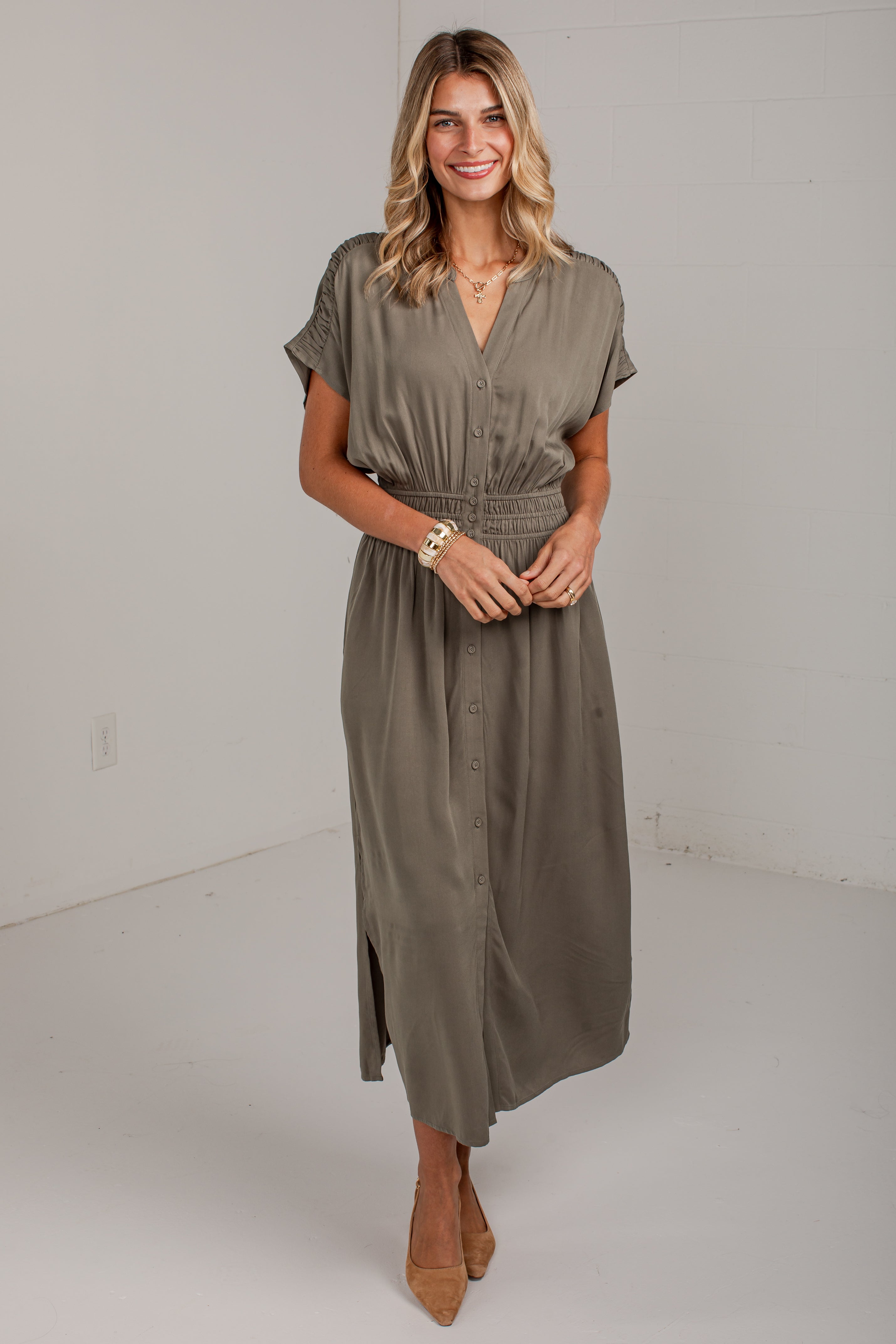 A woman modeling a short-sleeve button-front midi dress in a muted taupe-gray color, standing against a plain white studio wall.