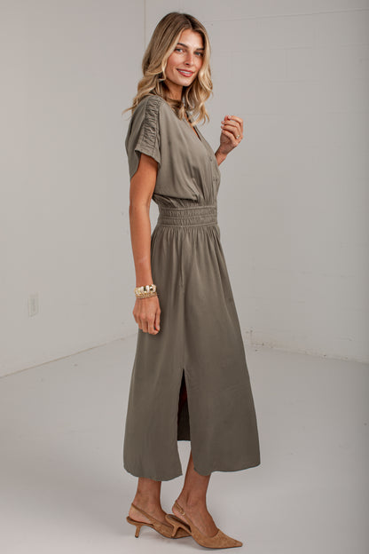A woman modeling a short-sleeve, knee-length olive green dress with a smocked waist and side slit, paired with tan heeled sandals, in a bright studio setting.