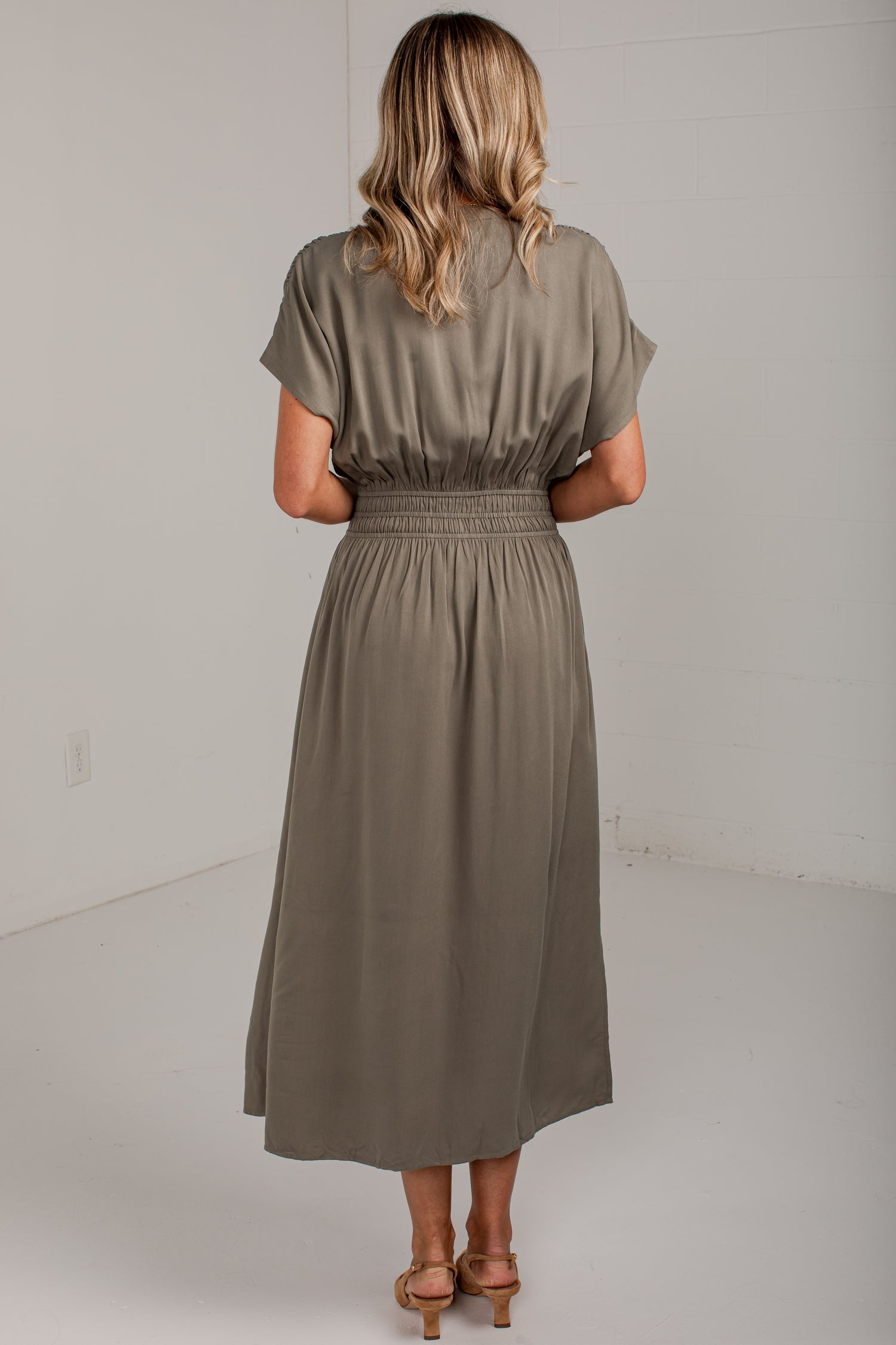 A woman viewed from behind wearing a short-sleeve, midi-length dress in a muted taupe or olive-brown shade. The dress has a gathered elastic waist and is shown against a plain white studio background.